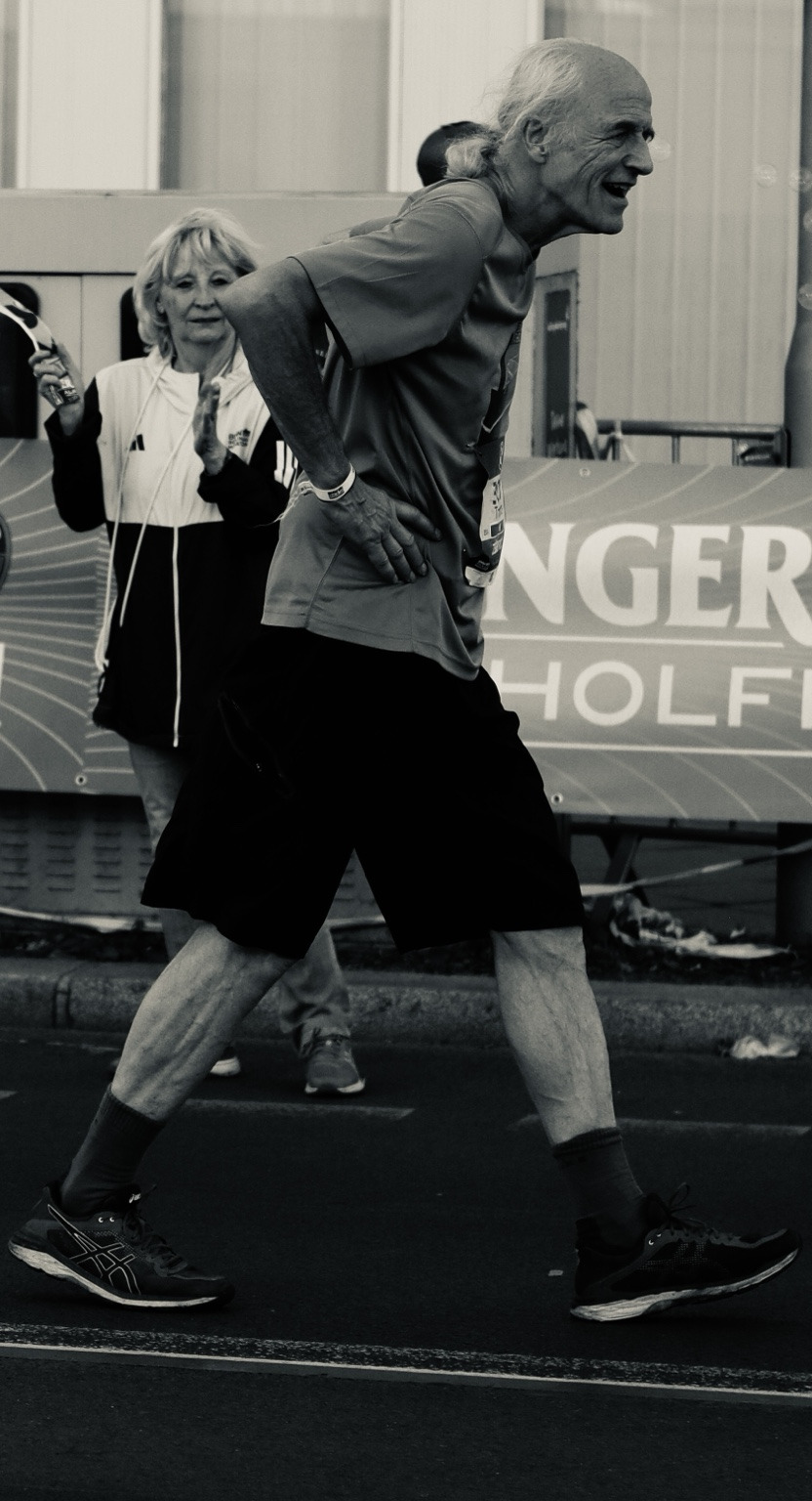 A runner strides forward on the Berlin Marathon course, photographed in black and white with spectators and event signage behind him.