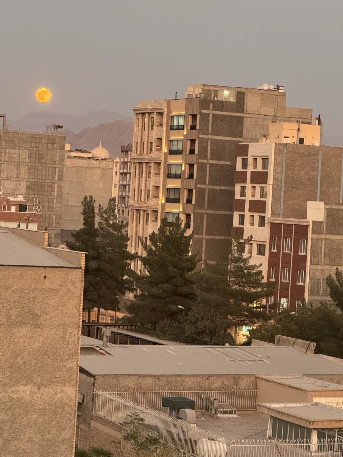ماه کامل بر فراز پشت‌بام‌های کرمان، با کوه‌ها و ساختمان‌های مسکونی در نور شامگاهی.